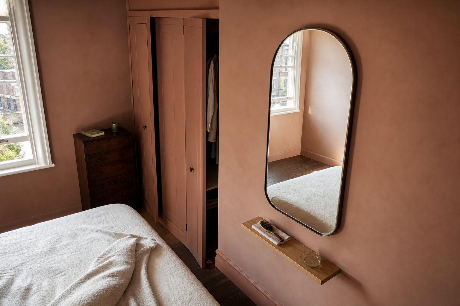 Compact bedroom with a tall arched smoked-bronze mirror beside a wardrobe, reflecting a bright window and pale wall